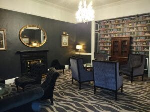 Picture of the inside of the room reserved for trans, intersex, and gender diverse conference attendees. It shows a cozy room with high backed arm chairs, green wallpaper on one size and wall paper made to look like a book shelf on the other. The carpet has a zebra print.