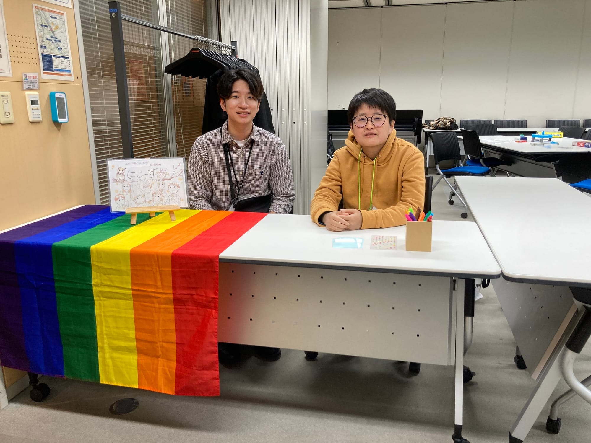 Picture of Mameta Endo and a colleague at a table for Picture of people at Niji-zu（24zzz) with a pride flag.