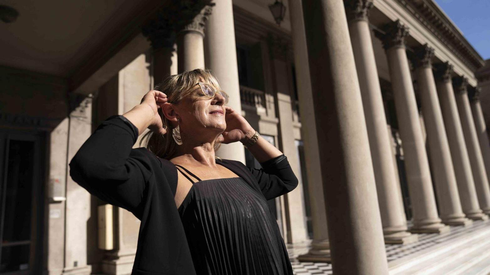 Picture of woman standing in front of building. She is wearing a black strapped shirt and coat. Her eyes are closed.