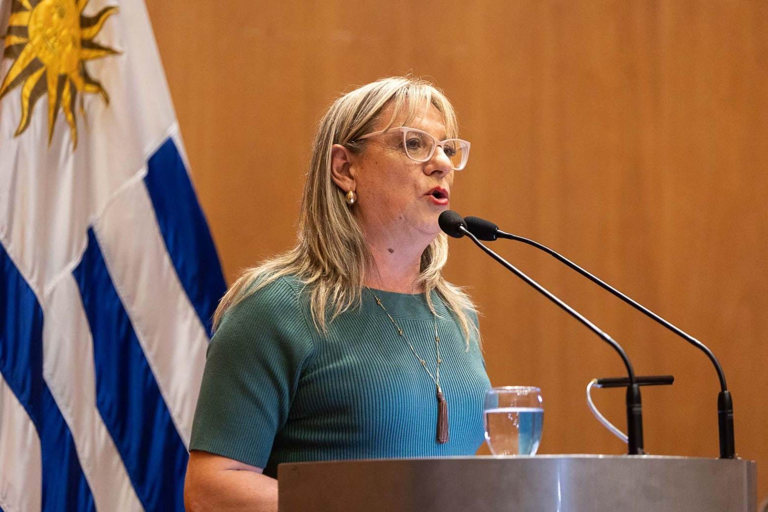 Picture of woman at lecturn. The Uruguayan flag is behind her and she wearing a blue shirt and speaking into two microphones.