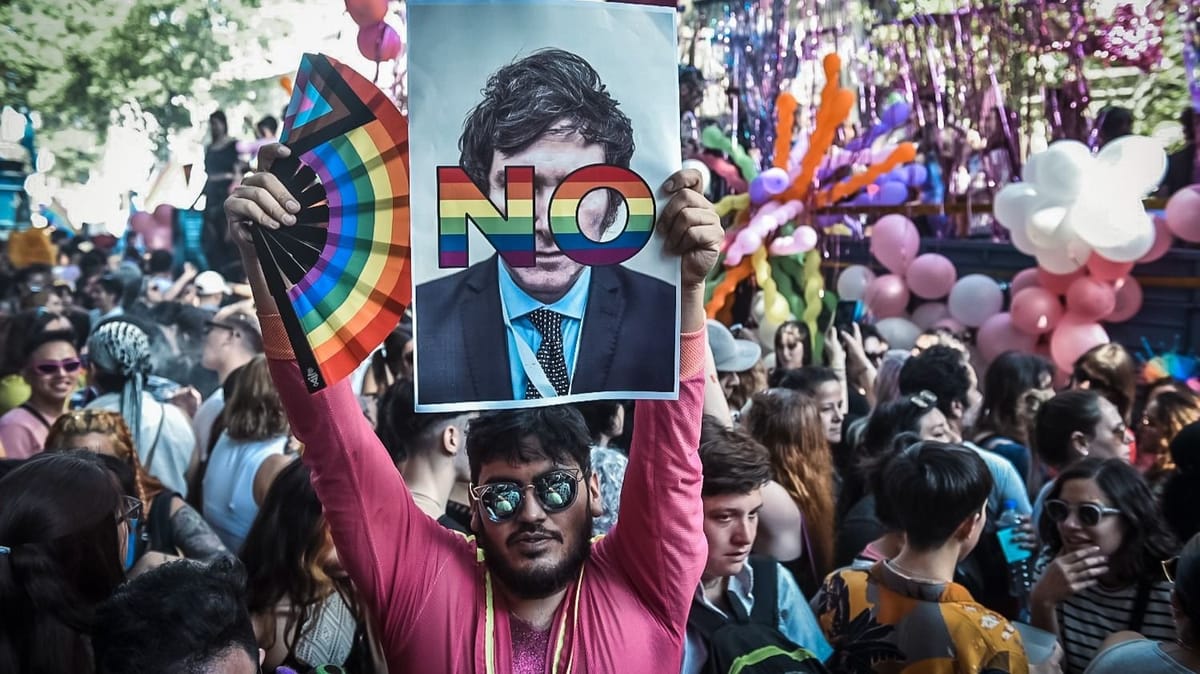 Person in large demonstration holding rainbow-coloured fan and picture of Javier Milei with NO on his face in rainbow.