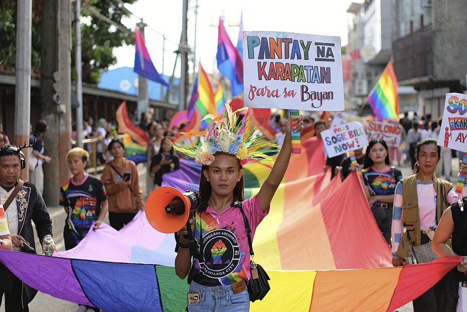 Image of a woman holding a sign that says Pantay na Karapatan para sa Bayan. She is standing in front of a large pride flag.