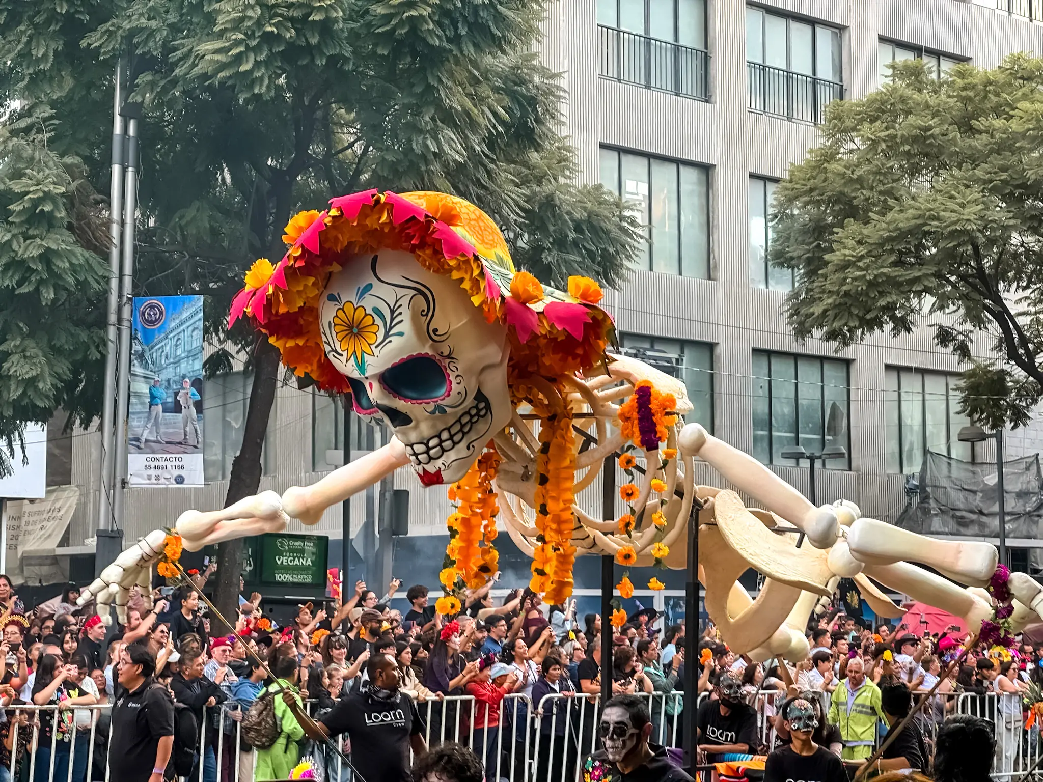Picture of a sculpture in a Day of the Dead parade. A Skeleton with a hat and flower necklace floats above the crowd.